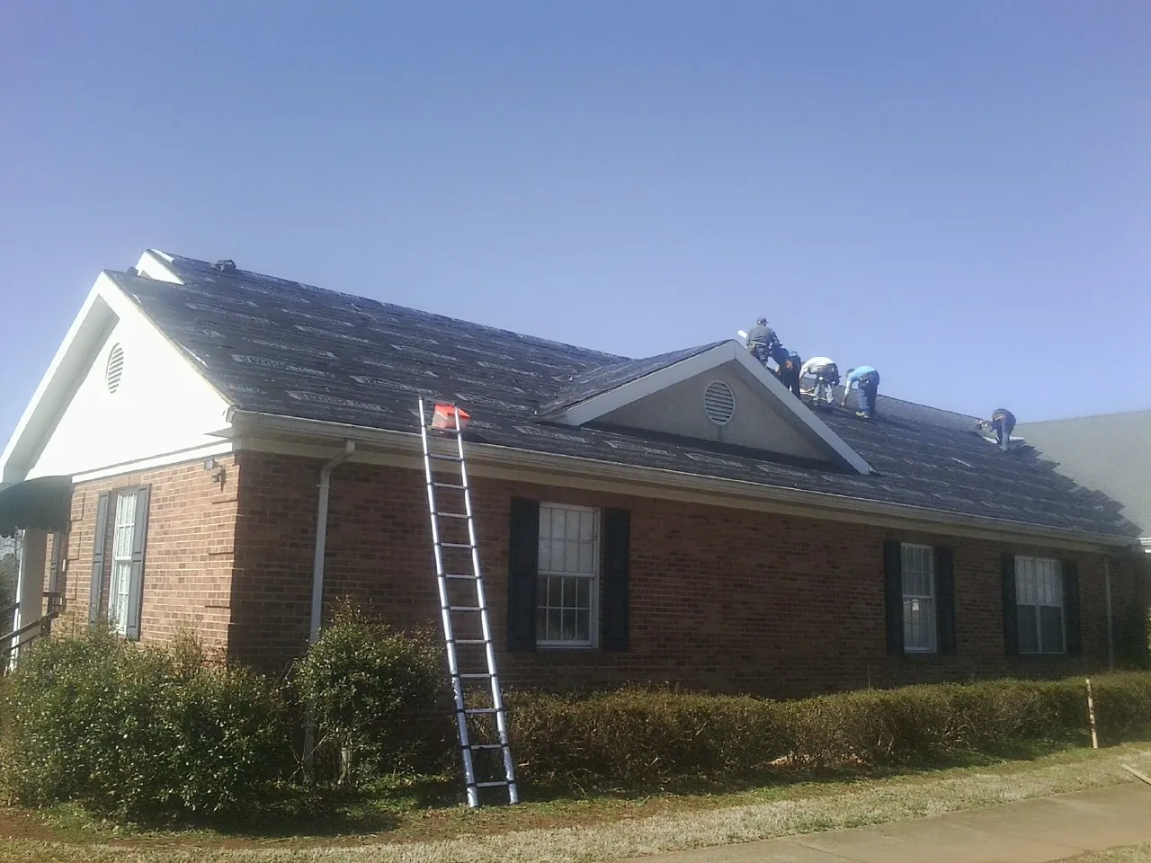 Crew tearing shingles off a brick ranch roof with a ladder set — Emergency Roof Repair work by our crew in Pleasant Garden