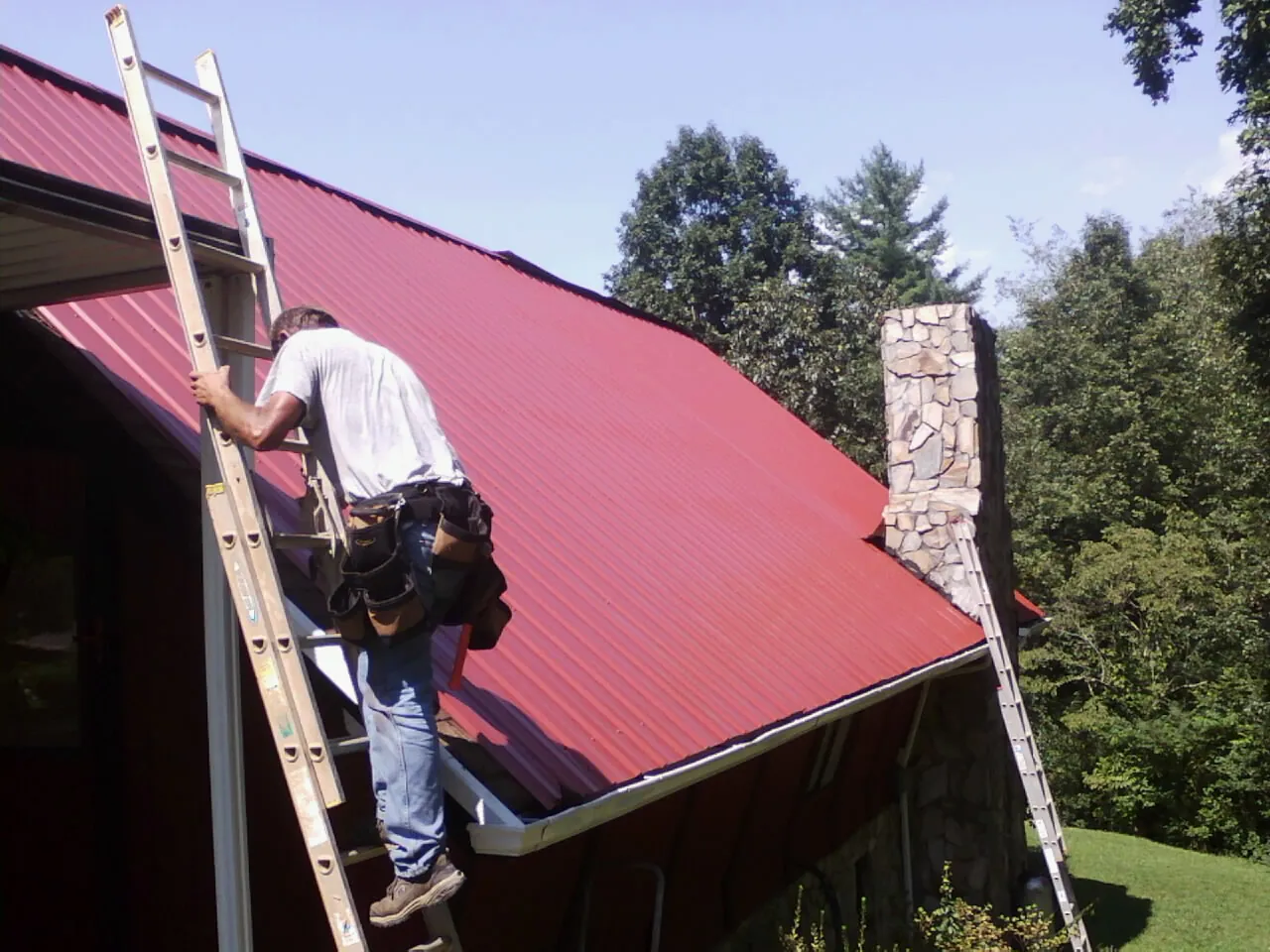 Roofer on a ladder beside a red metal roof and stone chimney — Soffit & Fascia Repair by Roofing Brothers in Pleasant Garden